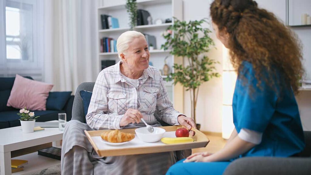 Older couple sharing a meal in a comfortable assisted living apartment while a caregiver offers discreet support