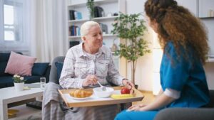 Older couple sharing a meal in a comfortable assisted living apartment while a caregiver offers discreet support