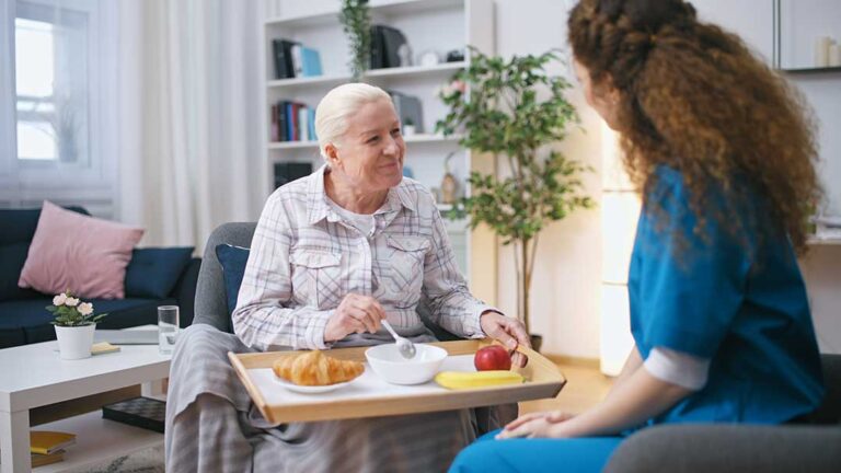 Older couple sharing a meal in a comfortable assisted living apartment while a caregiver offers discreet support